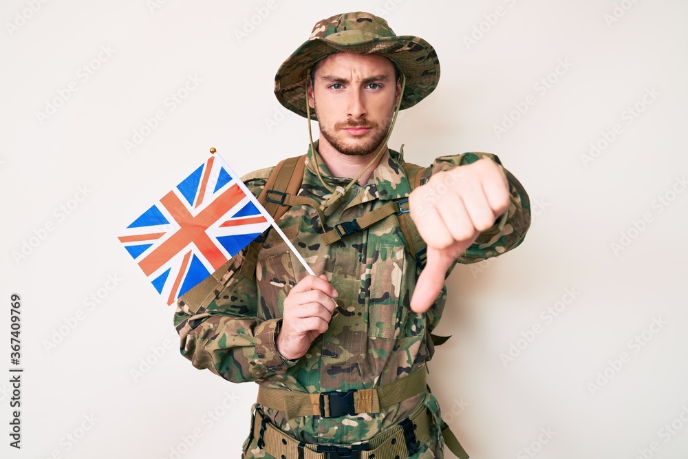 Fototapeta premium Young caucasian man wearing camouflage army uniform holding united kingdom flag with angry face, negative sign showing dislike with thumbs down, rejection concept