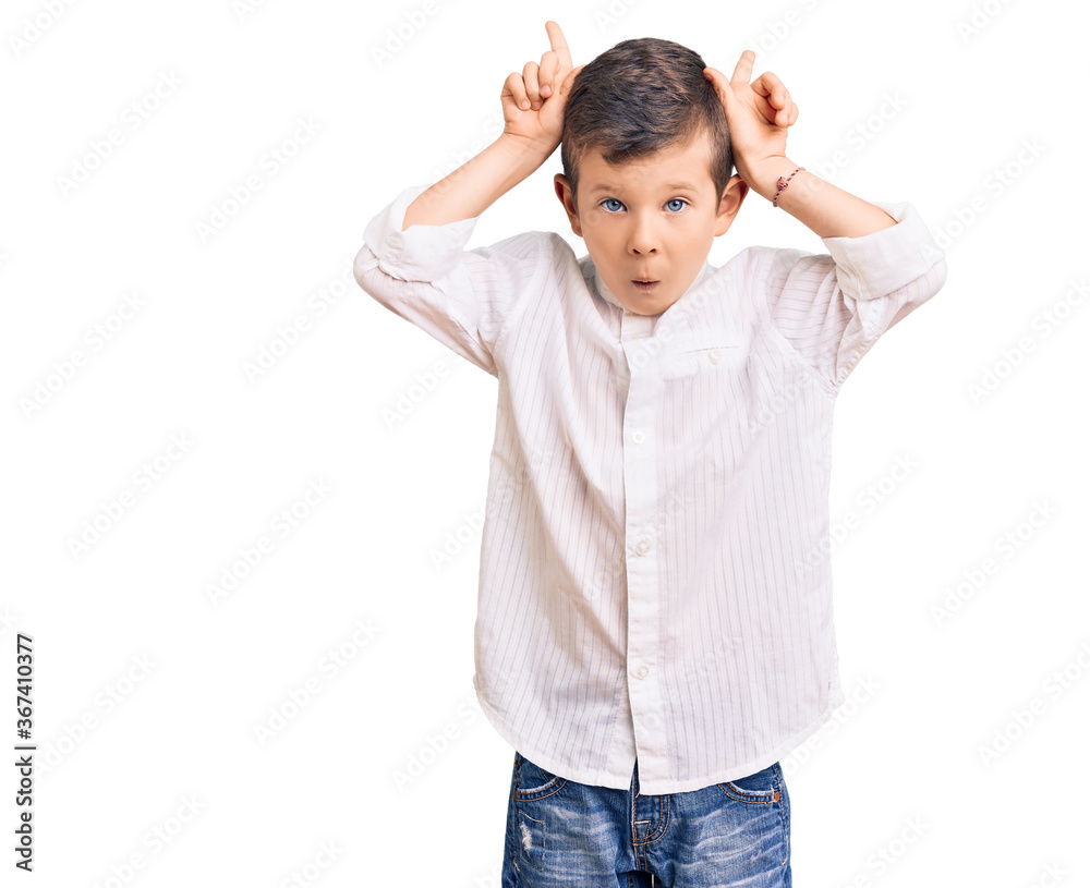 Cute blond kid wearing elegant shirt doing funny gesture with finger over head as bull horns Cute blond kid wearing elegant shirt doing funny gesture with finger over head as bull horns