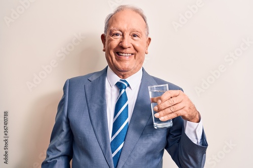 Senior handsome grey-haired businessman wearing suit drinking glass of water to refreshment looking positive and happy standing and smiling with a confident smile showing teeth © Krakenimages.com