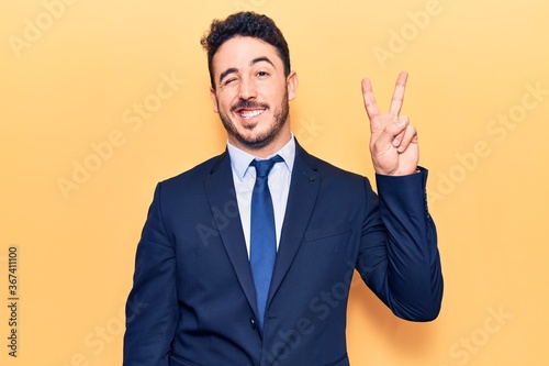 Young hispanic man wearing suit smiling with happy face winking at the camera doing victory sign. number two.
