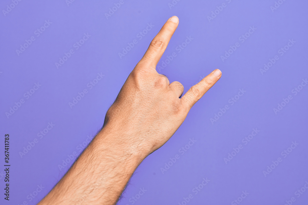 Hand of caucasian young man showing fingers over isolated purple background gesturing rock and roll symbol, showing obscene horns gesture
