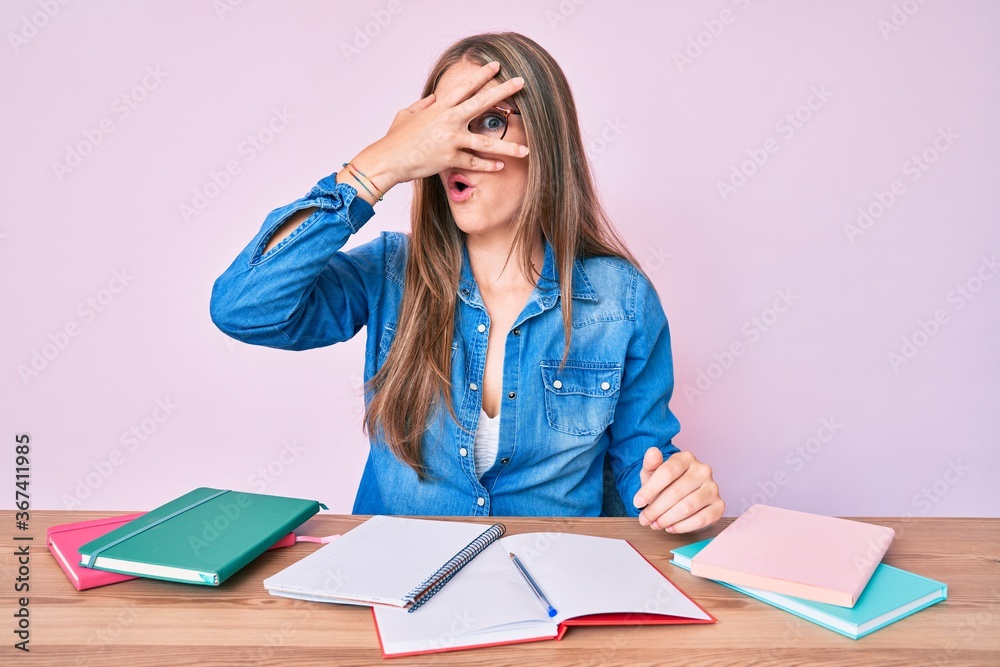 Young blonde girl studying sitting on the table peeking in shock covering face and eyes with hand, looking through fingers afraid Young blonde girl studying sitting on the table peeking in shock covering face and eyes with hand, looking through fingers afraid