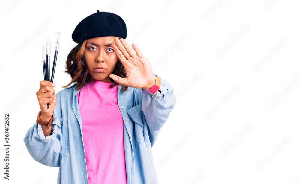 Young beautiful mixed race woman holding paintbrushes with open hand doing stop sign with serious and confident expression, defense gesture Young beautiful mixed race woman holding paintbrushes with open hand doing stop sign with serious and confident expression, defense gesture