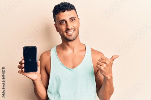 Young hispanic man holding smartphone showing screen smiling happy and positive, thumb up doing excellent and approval sign