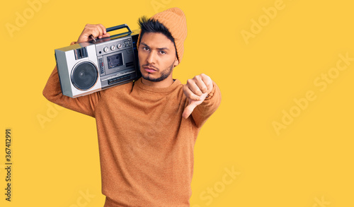 Handsome latin american young man holding boombox, listening to music looking unhappy and angry showing rejection and negative with thumbs down gesture. bad expression.
