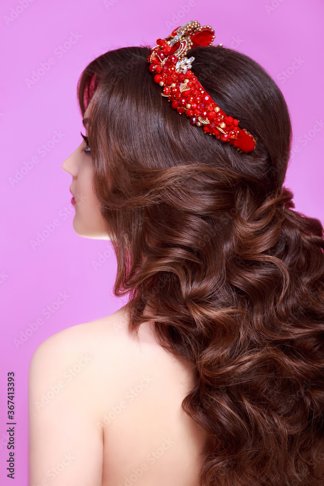 Naklejka premium Beautiful brunette woman with long Curly hair wearing crown decorated gemstones. Fashion makeup and hairstyle. Portrait of young gorgeous bride. Isolated on pink background