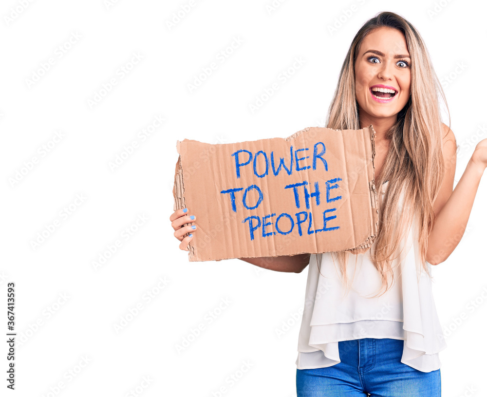 Young beautiful blonde woman holding power to the people cardboard banner celebrating victory with happy smile and winner expression with raised hands Young beautiful blonde woman holding power to the people cardboard banner celebrating victory with happy smile and winner expression with raised hands