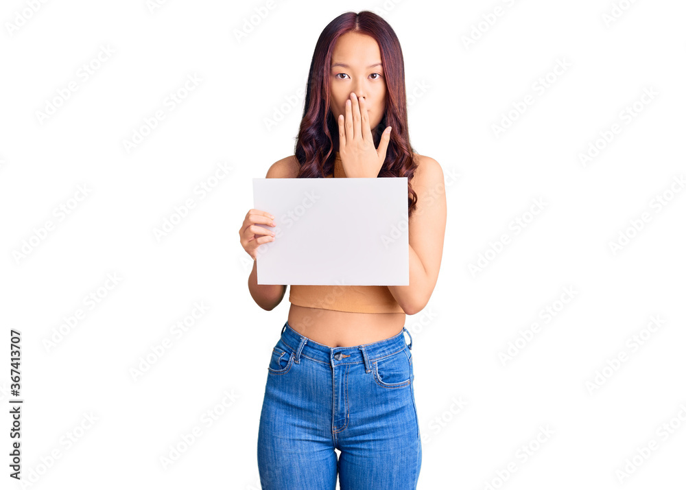 Young beautiful chinese girl holding paper banner with blank space covering mouth with hand, shocked and afraid for mistake. surprised expression Young beautiful chinese girl holding paper banner with blank space covering mouth with hand, shocked and afraid for mistake. surprised expression