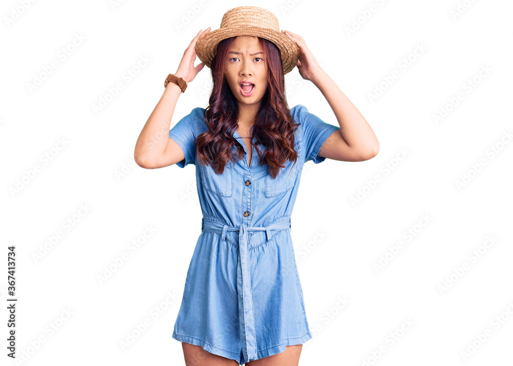 Young beautiful chinese girl wearing summer hat crazy and scared with hands on head, afraid and surprised of shock with open mouth Young beautiful chinese girl wearing summer hat crazy and scared with hands on head, afraid and surprised of shock with open mouth