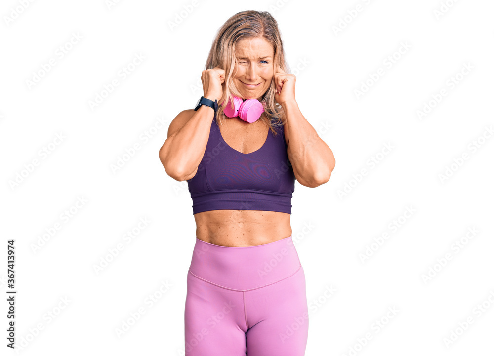 Middle age fit blonde woman wearing gym clothes and using headphones covering ears with fingers with annoyed expression for the noise of loud music. deaf concept.