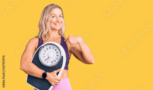Middle age fit blonde woman wearing sports clothes holding weighing machine pointing finger to one self smiling happy and proud