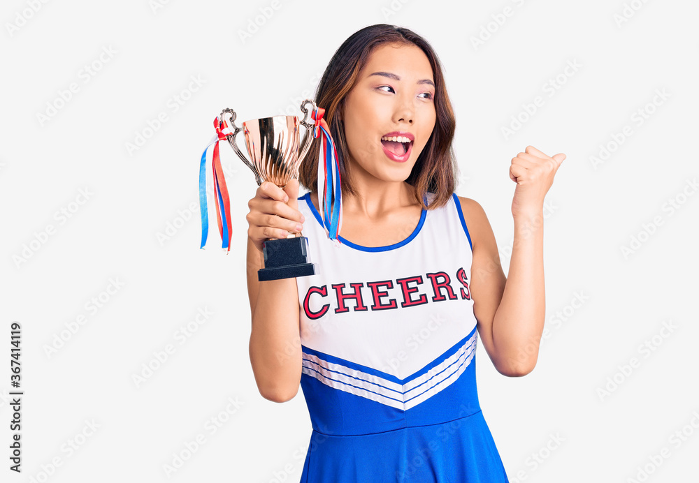 Youn beautiful asian girl wearing cheerleader uniform holding champion trophy pointing thumb up to the side smiling happy with open mouth Youn beautiful asian girl wearing cheerleader uniform holding champion trophy pointing thumb up to the side smiling happy with open mouth