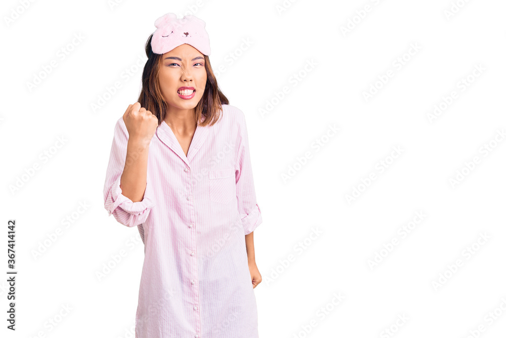 Young beautiful chinese girl wearing sleep mask and pajama angry and mad raising fist frustrated and furious while shouting with anger. rage and aggressive concept. Young beautiful chinese girl wearing sleep mask and pajama angry and mad raising fist frustrated and furious while shouting with anger. rage and aggressive concept.