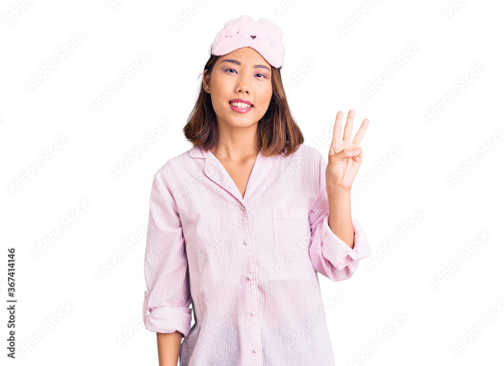 Young beautiful chinese girl wearing sleep mask and pajama showing and pointing up with fingers number three while smiling confident and happy. Young beautiful chinese girl wearing sleep mask and pajama showing and pointing up with fingers number three while smiling confident and happy.