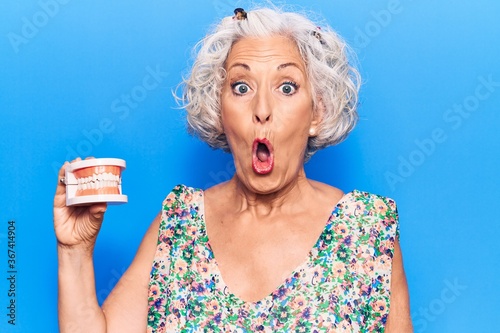 Senior grey-haired woman holding denture scared and amazed with open mouth for surprise, disbelief face © Krakenimages.com