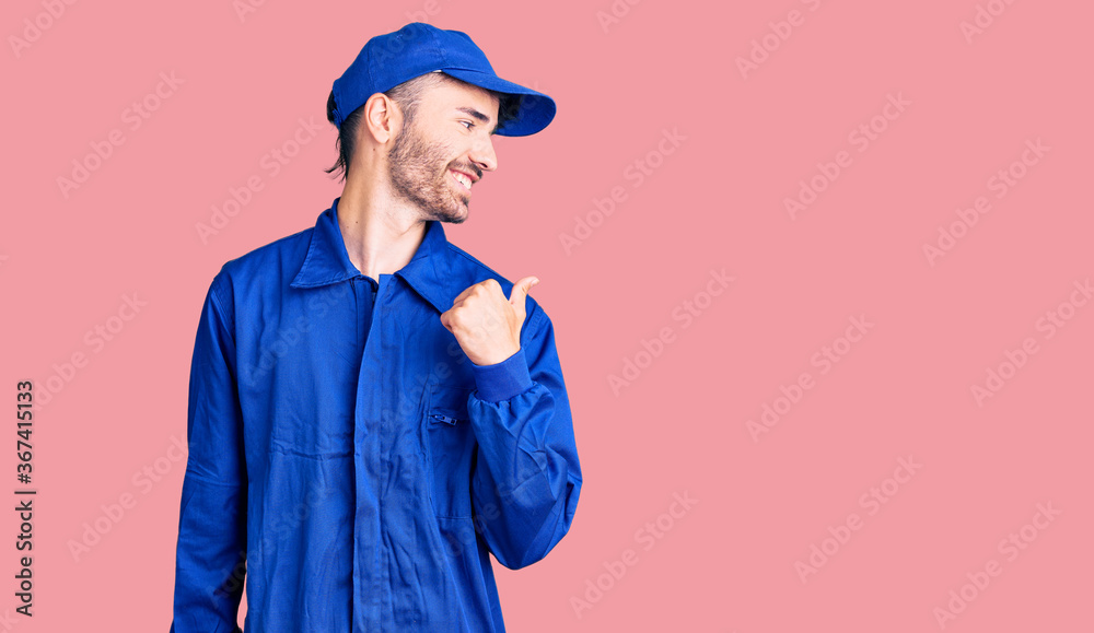 Young hispanic man wearing painter uniform smiling with happy face looking and pointing to the side with thumb up. Young hispanic man wearing painter uniform smiling with happy face looking and pointing to the side with thumb up.