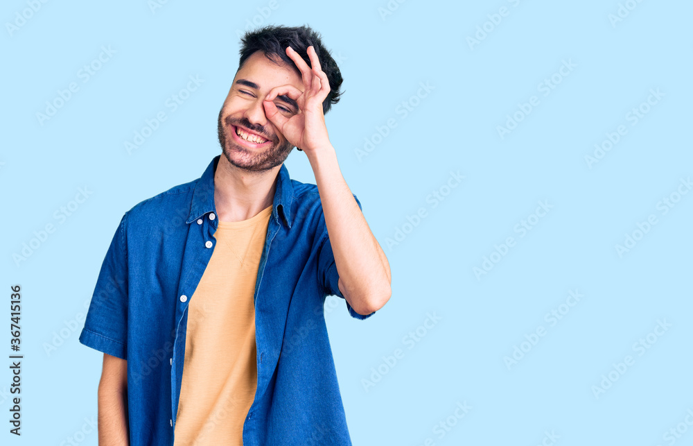 Young hispanic man wearing casual clothes doing ok gesture with hand smiling, eye looking through fingers with happy face. Young hispanic man wearing casual clothes doing ok gesture with hand smiling, eye looking through fingers with happy face.