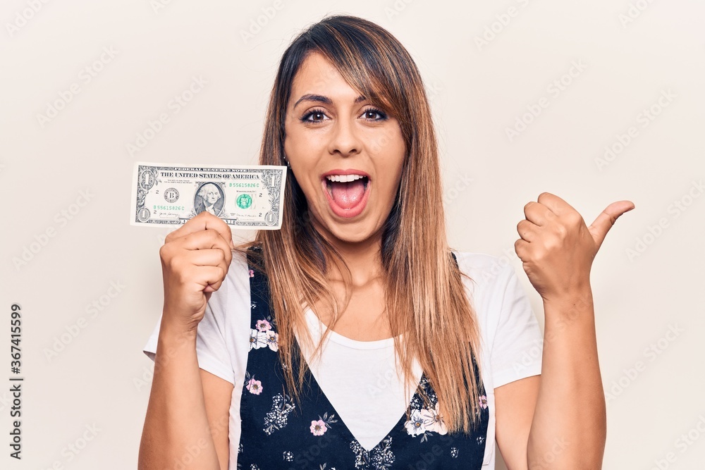 Young beautiful woman holding one dollar pointing thumb up to the side smiling happy with open mouth