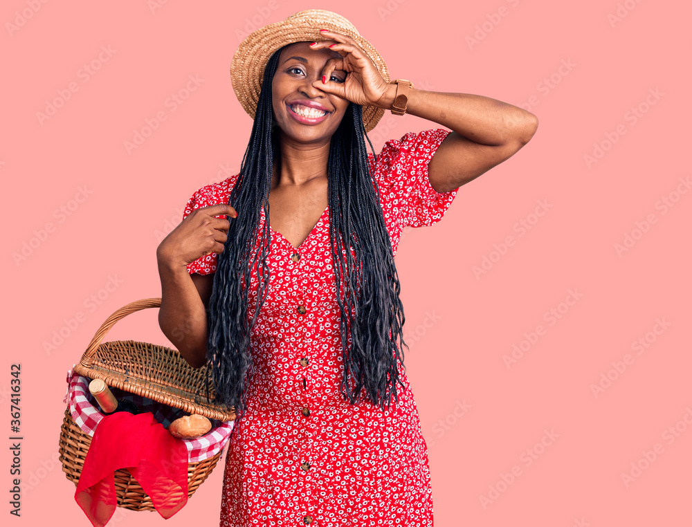 Young african american woman wearing summer hat and holding picnic wicker basket with bread smiling happy doing ok sign with hand on eye looking through fingers Young african american woman wearing summer hat and holding picnic wicker basket with bread smiling happy doing ok sign with hand on eye looking through fingers