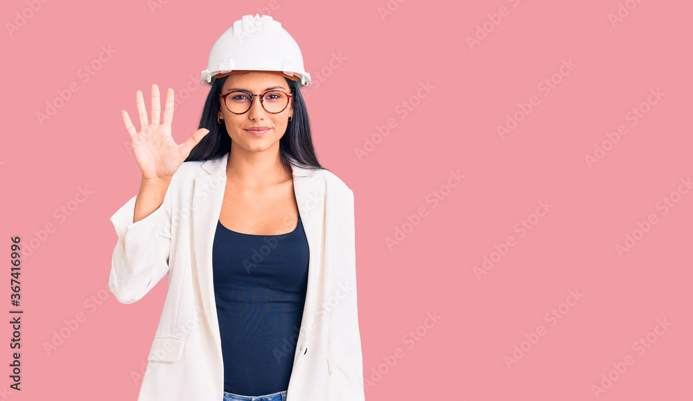 Young beautiful latin girl wearing architect hardhat and glasses showing and pointing up with fingers number five while smiling confident and happy. Young beautiful latin girl wearing architect hardhat and glasses showing and pointing up with fingers number five while smiling confident and happy.