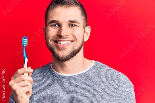 Young handsome man holding toothbrush looking positive and happy standing and smiling with a confident smile showing teeth