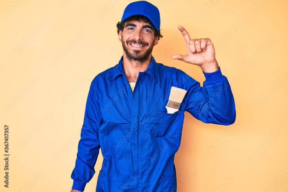Handsome young man with curly hair and bear wearing builder jumpsuit uniform smiling and confident gesturing with hand doing small size sign with fingers looking and the camera. measure concept.