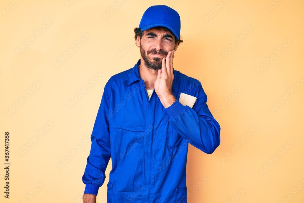 Handsome young man with curly hair and bear wearing builder jumpsuit uniform touching mouth with hand with painful expression because of toothache or dental illness on teeth. dentist