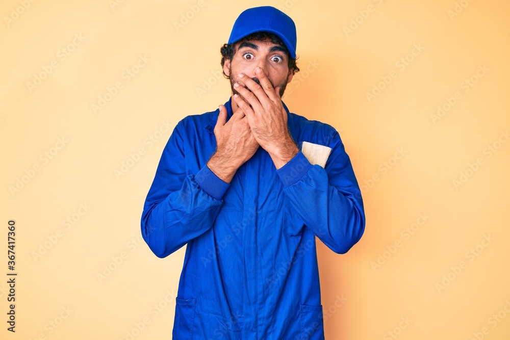 Handsome young man with curly hair and bear wearing builder jumpsuit uniform shocked covering mouth with hands for mistake. secret concept.