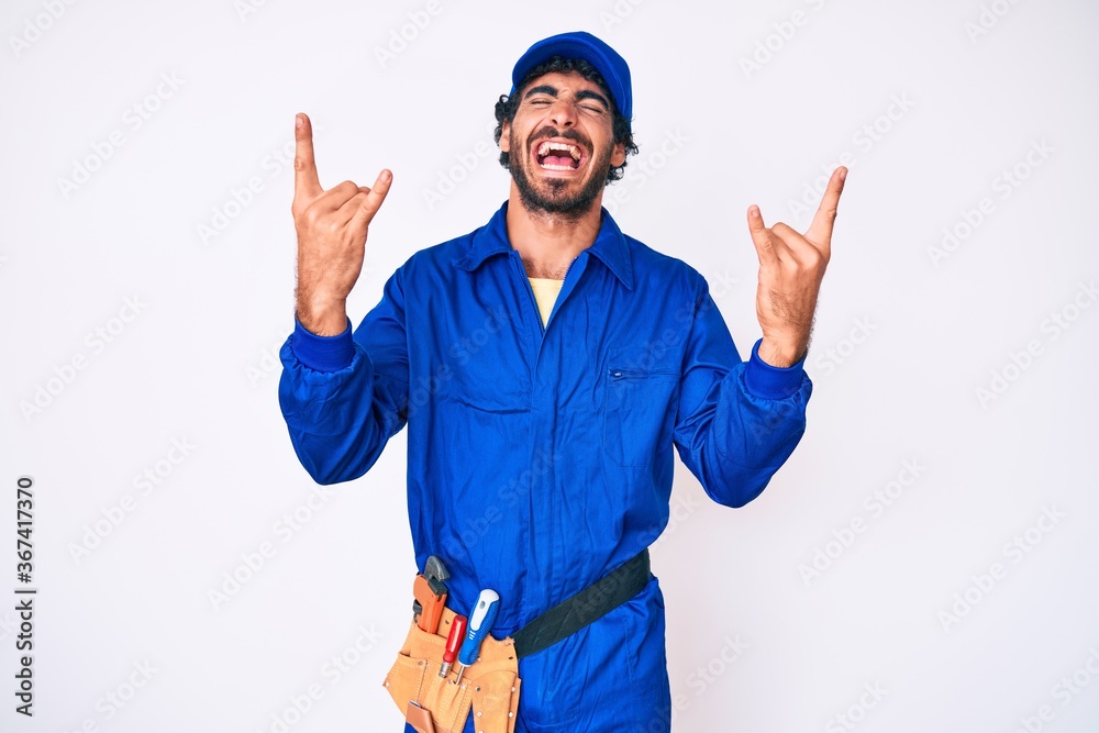 Handsome young man with curly hair and bear weaing handyman uniform shouting with crazy expression doing rock symbol with hands up. music star. heavy music concept.