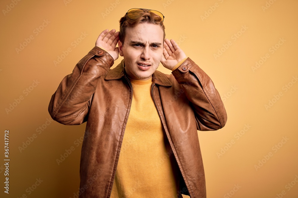 Young handsome redhead man wearing casual leather jacket over isolated yellow background Trying to hear both hands on ear gesture, curious for gossip. Hearing problem, deaf Young handsome redhead man wearing casual leather jacket over isolated yellow background Trying to hear both hands on ear gesture, curious for gossip. Hearing problem, deaf