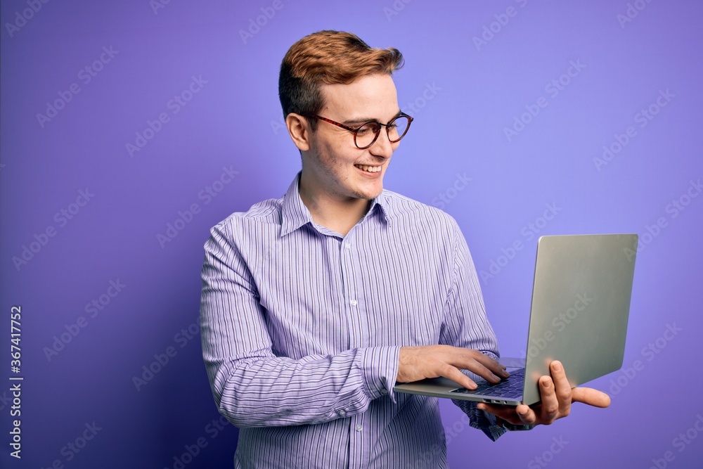 Young handsome redhead businessman working using laptop over purple background with a happy face standing and smiling with a confident smile showing teeth Young handsome redhead businessman working using laptop over purple background with a happy face standing and smiling with a confident smile showing teeth