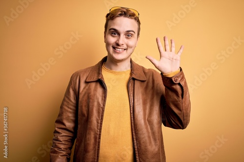 Young handsome redhead man wearing casual leather jacket over isolated yellow background showing and pointing up with fingers number five while smiling confident and happy. © Krakenimages.com
