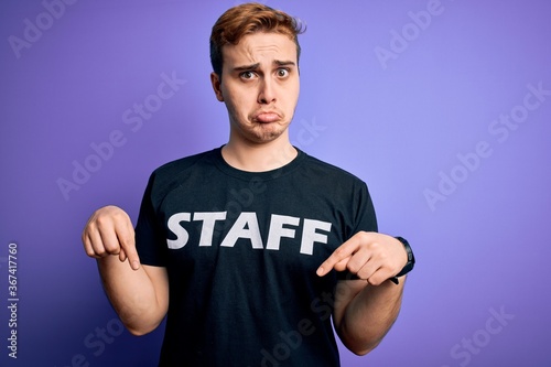 Young handsome redhead worker man wearing staff t-shirt uniform over purple background Pointing down looking sad and upset, indicating direction with fingers, unhappy and depressed. © Krakenimages.com