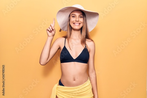 Young beautiful girl wearing bikini and hat smiling with an idea or question pointing finger up with happy face  number one