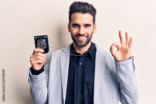 Young handsome man with beard holding police badge doing ok sign with fingers, smiling friendly gesturing excellent symbol © Krakenimages.com
