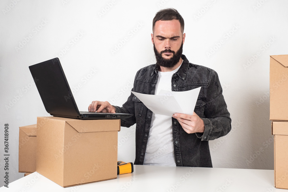 A serious man next to a laptop. A man is holding documents in his hands. A man in a warehouse next to cardboard boxes. Acceptance of goods to the warehouse. A serious man next to a laptop. A man is holding documents in his hands. A man in a warehouse next to cardboard boxes. Acceptance of goods to the warehouse.