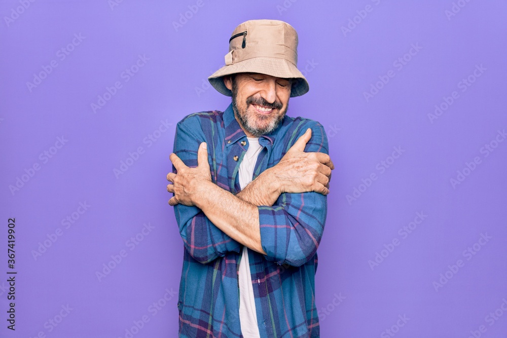 Middle age handsome man wearing casual shirt and hat over isolated purple background hugging oneself happy and positive, smiling confident. Self love and self care Middle age handsome man wearing casual shirt and hat over isolated purple background hugging oneself happy and positive, smiling confident. Self love and self care