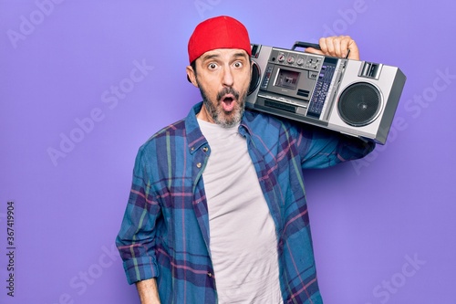 Middle age man wearing cap listening vintage boombox over isolated purple background scared and amazed with open mouth for surprise, disbelief face