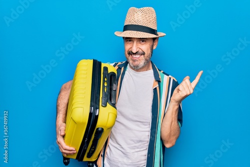 Middle age handsome tourist man on vacation holding cabin bag over isolated blue background smiling happy pointing with hand and finger to the side © Krakenimages.com