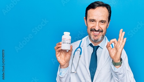 Middle age handsome doctor man wearing stethoscope holding bottle of pills doing ok sign with fingers, smiling friendly gesturing excellent symbol