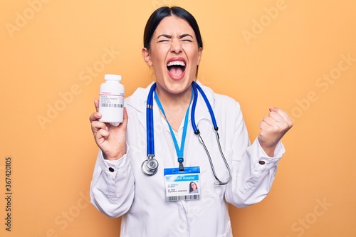 Young doctor woman wearing stethoscope holding bottle of pills over yellow background screaming proud  celebrating victory and success very excited with raised arm