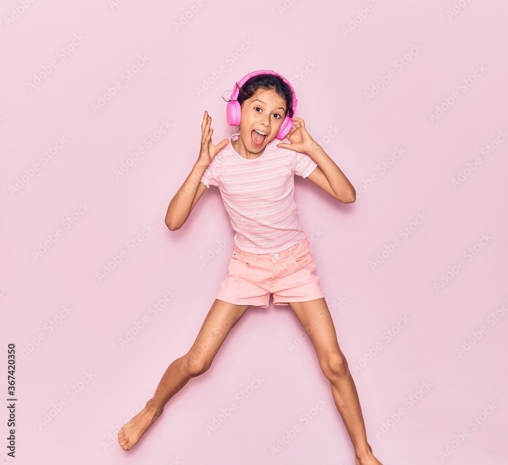 Adorable hispanic child girl smiling happy. Jumping with smile on face listening to music using pink headphones over isolated background Adorable hispanic child girl smiling happy. Jumping with smile on face listening to music using pink headphones over isolated background