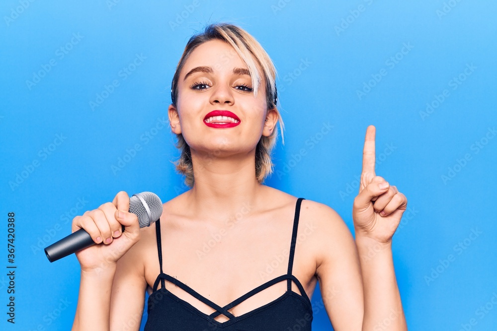 Young beautiful blonde woman singing song using microphone smiling with an idea or question pointing finger with happy face, number one