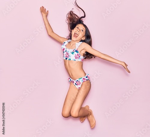 Adorable hispanic child girl on vacation wearing bikini surprised with open mouth. Jumping over isolated pink background