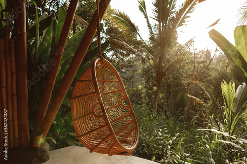 Wicker rattan hanging chair on wooden bamboo terrace in the jungle, nature view. Rattan lounge hanging chair at the balcony with green nature background #367424187