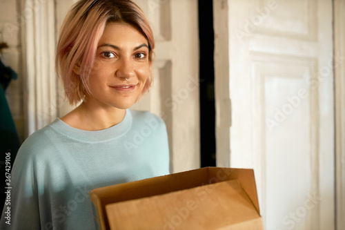 Woman get online goods from the shop. Girl stay with box at home and look into the camera. Shopping in store covid time. Happy lady take delivery food.