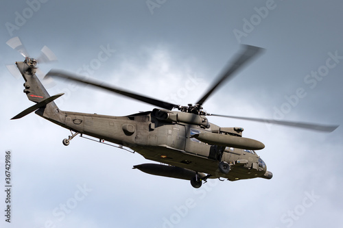 Army helicopter with large fuel tanks flying past dark clouds.