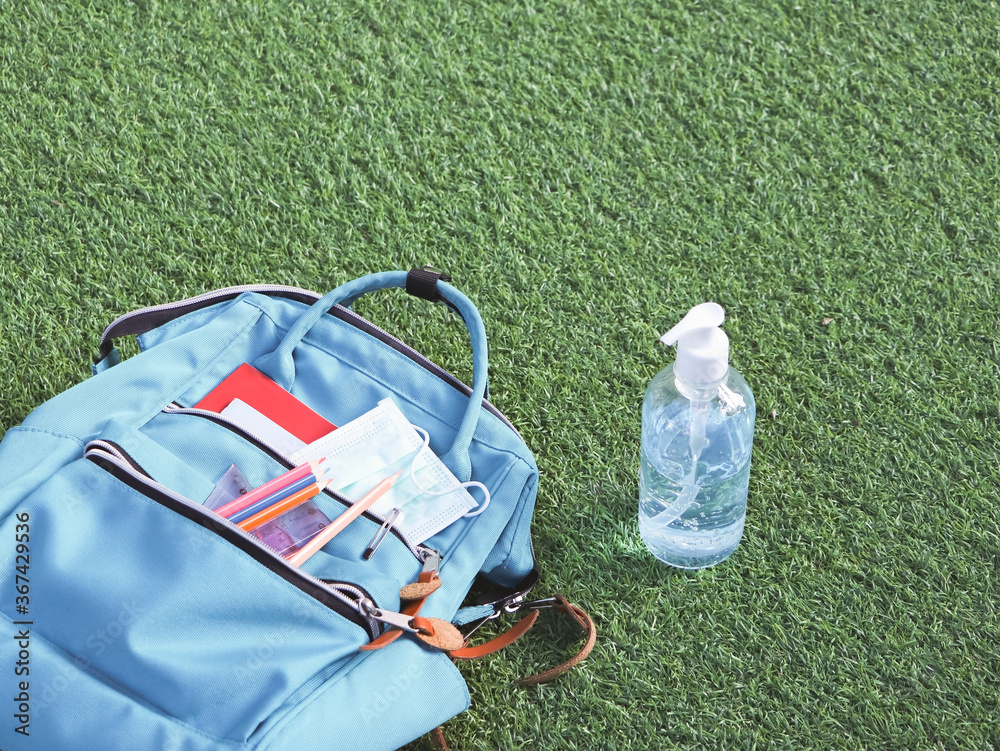 COVID-19 prevention while going back  to school  and new normal  concept. Top view of backpack with school supplies , surgical mask and alcohol sanitizer gel on green grass