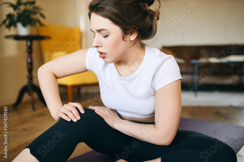 Portrait of unhappy sporty girl grabbing knee, trying to massage aching area, unable to do yoga because of sport injury, feeling pain. Young female beginner doing wrong exercise without trainer © Anatoliy Karlyuk