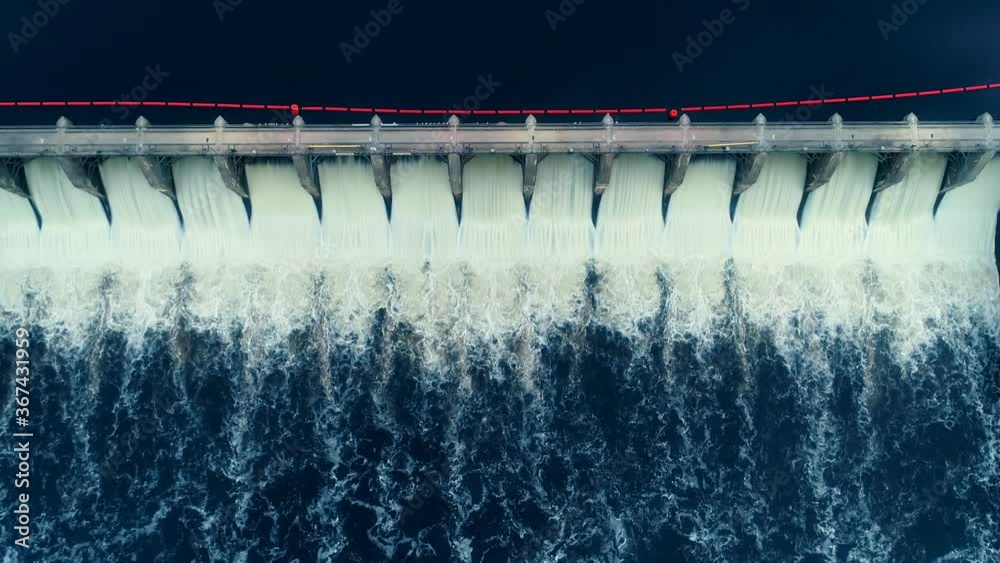 Aerial flyover shot of a dam in full release mode. The release creates this white ink like texture on the surface of the dark blue water.